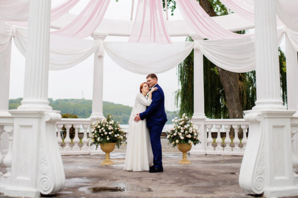 Wedding couple under large arch with pink material decoration with flowers.