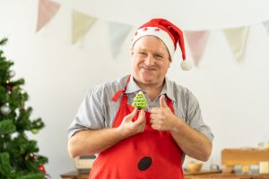 Happy senior man in kitchen apron decorating Christmas gingerbread cookies on Christmas. Chef work in the cuisine. Cook in uniform. Merry Christmas and Happy New Year. Cooking and Holiday concept